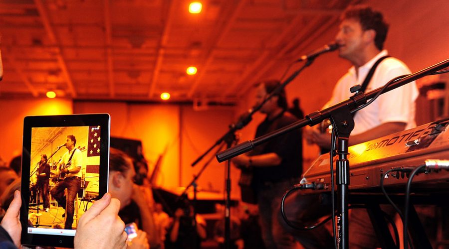 Musicians performing live in Austin, with an individual recording the show on a computer tablet